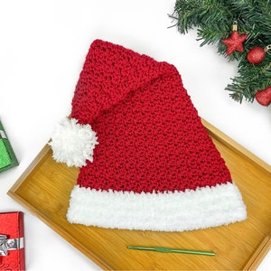Könnte beinhalten: Ein roter gehäkelter Weihnachtsmannhut mit weißem, flauschigem Besatz und einem weißen Bommel. Der Hut wird auf einem Holztablett präsentiert, mit Weihnachtsdekorationen im Hintergrund. Ein grüner Häkelhaken ist ebenfalls sichtbar.