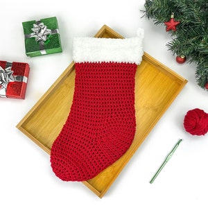 May include: Red and white crocheted Christmas stocking with a white fur cuff, sitting on a wooden tray with a ball of red yarn and a green crochet hook.