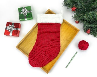 Puede incluir: Calcetín de Navidad de crochet rojo y blanco con un borde de piel blanca, sentado en una bandeja de madera con una bola de hilo rojo y un gancho de crochet verde.