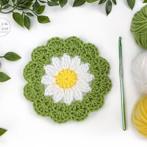 May include: A green crochet daisy flower with a white centre and yellow centre. The flower is surrounded by green yarn and a crochet hook.