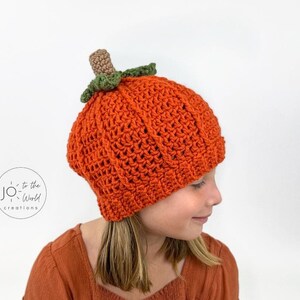 K&ouml;nnte beinhalten: Eine geh&auml;kelte K&uuml;rbis-M&uuml;tze in Orange mit einem gr&uuml;nen Stiel und Bl&auml;ttern. Die M&uuml;tze wird von einem Kind mit langen braunen Haaren getragen.