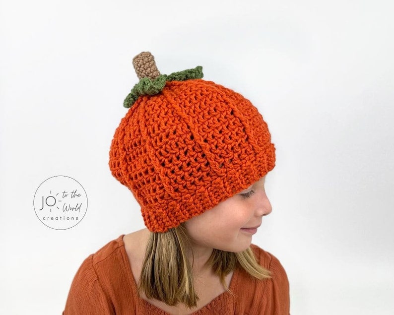 K&ouml;nnte beinhalten: Eine geh&auml;kelte K&uuml;rbis-M&uuml;tze in Orange mit einem gr&uuml;nen Stiel und Bl&auml;ttern. Die M&uuml;tze wird von einem Kind mit langen braunen Haaren getragen.