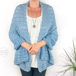 May include: A light blue, open-weave crochet cardigan. The cardigan has a draped design with three-quarter sleeves. The garment is worn over a white top and a silver necklace. The cardigan is a soft, airy, and comfortable garment.