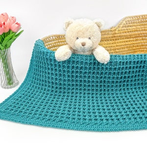 Puede incluir: Una manta de bebé de crochet azul turquesa con un patrón de punto de waffle. Un osito de peluche asoma por detrás de la manta.