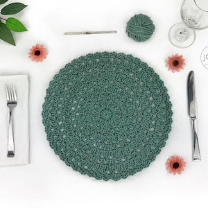 May include: A teal crochet placemat with a circular pattern, displayed on a white surface. The image features a fork, knife, napkin, and a wine glass. Decorative flowers and a ball of yarn are also present, creating a handcrafted aesthetic.