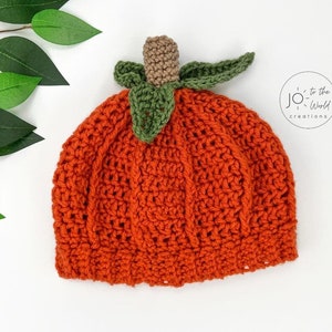 K&ouml;nnte beinhalten: Orangefarbener H&auml;kelhut in Form eines K&uuml;rbisses mit gr&uuml;nen Bl&auml;ttern und einem braunen Stiel. Der Hut ist mit einem strukturierten Stich gearbeitet und hat eine abgerundete Form.