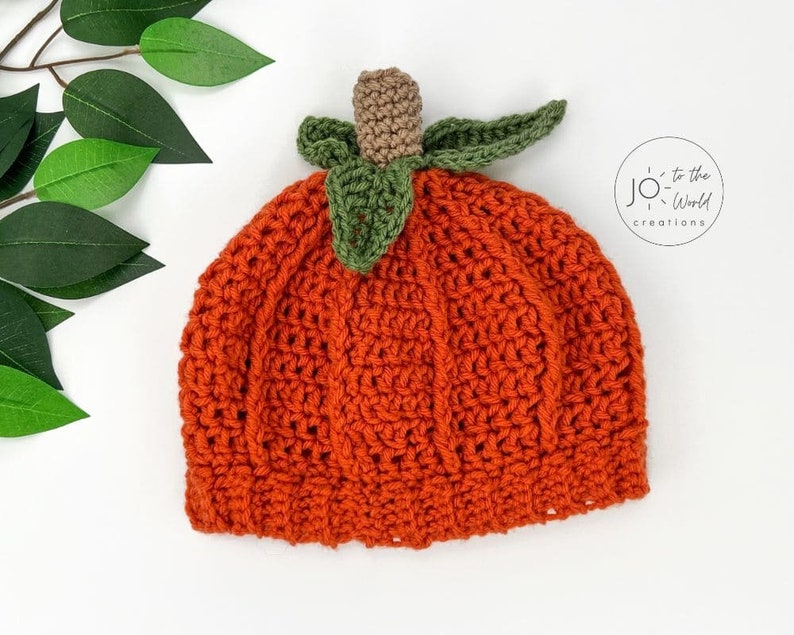 K&ouml;nnte beinhalten: Orangefarbener H&auml;kelhut in Form eines K&uuml;rbisses mit gr&uuml;nen Bl&auml;ttern und einem braunen Stiel. Der Hut ist mit einem strukturierten Stich gearbeitet und hat eine abgerundete Form.