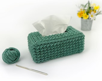 May include: A green crocheted tissue box cover with a white tissue box inside. A ball of green yarn and a crochet hook are also in the image.