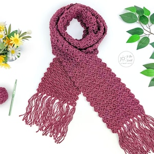 May include: A deep magenta crochet scarf featuring a textured stitch pattern and long fringe at both ends. The scarf is displayed alongside a matching ball of yarn, a green crochet hook, a small bouquet of yellow and white flowers, and green leaves.