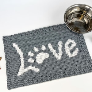 Op de afbeelding: Grijze gehaakte hondenvoedingsmat met een witte pootprint en het woord "Love" in wit.