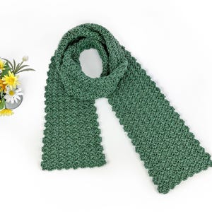 May include: A green, hand-knitted scarf with a textured pattern. The scarf is folded over itself, creating a loop. The scarf is made of a soft, textured yarn. A small bouquet of yellow and white flowers sits to the left of the scarf.