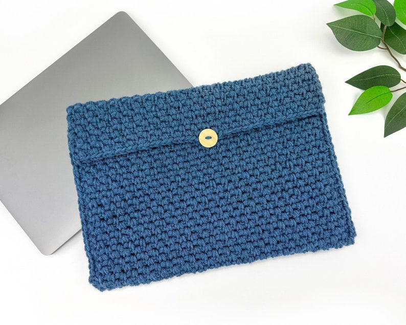May include: A blue crocheted laptop sleeve with a wooden button closure. The sleeve is lying on a white surface next to a silver laptop.