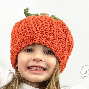 K&ouml;nnte beinhalten: Ein Kind tr&auml;gt eine geh&auml;kelte K&uuml;rbis-M&uuml;tze. Die M&uuml;tze ist orange mit gr&uuml;nen Bl&auml;ttern und einem braunen Stiel. Das Kind l&auml;chelt und schaut in die Kamera.