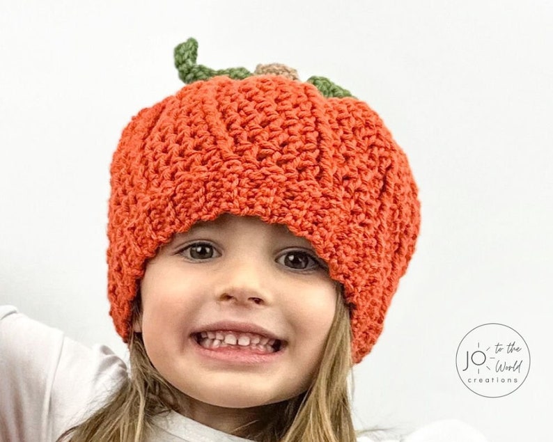 K&ouml;nnte beinhalten: Ein Kind tr&auml;gt eine geh&auml;kelte K&uuml;rbis-M&uuml;tze. Die M&uuml;tze ist orange mit gr&uuml;nen Bl&auml;ttern und einem braunen Stiel. Das Kind l&auml;chelt und schaut in die Kamera.