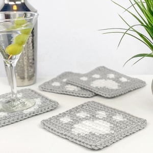 May include: Four gray crocheted coasters with white paw prints. The coasters are on a white surface next to a martini glass and a potted plant.