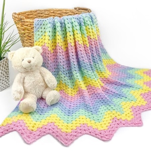 Op de afbeelding: Een pastelkleurige regenboog gehaakte babydeken met een chevron-patroon. De deken is gedrapeerd over een rieten mand en een knuffelbeer.