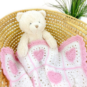 Pode incluir: Um urso de pelúcia creme repousa numa cesta de vime, sob um cobertor de crochê branco e rosa. O cobertor apresenta aplicações de coração rosa e uma borda ondulada. A cesta é feita de material tecido castanho claro.