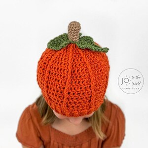 K&ouml;nnte beinhalten: Eine geh&auml;kelte K&uuml;rbis-M&uuml;tze in Orange mit einem gr&uuml;nen Stiel und Bl&auml;ttern. Die M&uuml;tze wird von einer Person mit braunen Haaren getragen.