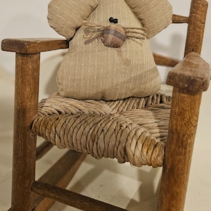 May include: A small, brown rocking chair with a woven straw seat. A brown fabric mouse with black eyes and a brown nose sits on the seat.