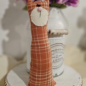 May include: A handmade orange and white plaid cat doll with a white felt face and black eyes. The doll is sitting on a white wooden tiered stand with a beaded edge.