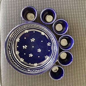 May include: A dark blue ceramic plate and matching small cups. The plate has white floral and wavy line designs. The cups are arranged around the plate. The set is likely for serving appetizers or small portions.