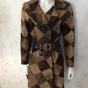 May include: A brown and tan patchwork coat with a double-breasted design. The coat features a black belt with a gold buckle and is displayed on a mannequin. The coat has a diamond pattern and is made of a suede-like material.