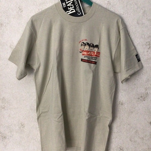 May include: A beige t-shirt with a Beatles graphic and the text "The Beatles British Invasion Pop Concert American Tour 1964". A black tag with "The Beatles Collectors Series" is attached to the collar.
