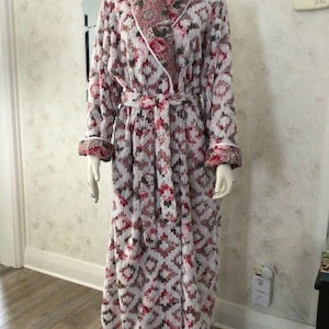 May include: A long, floral-patterned robe with a shawl collar and matching belt. The robe features a white background with a repeating pattern of red and pink flowers and geometric shapes. The cuffs and collar are trimmed with a floral print.