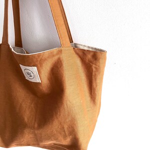 May include: A solid tobacco-colored tote bag featuring two long shoulder straps and a white rectangular fabric label on the front. The label contains a circular logo with black text and a small graphic element. The interior lining is off-white.