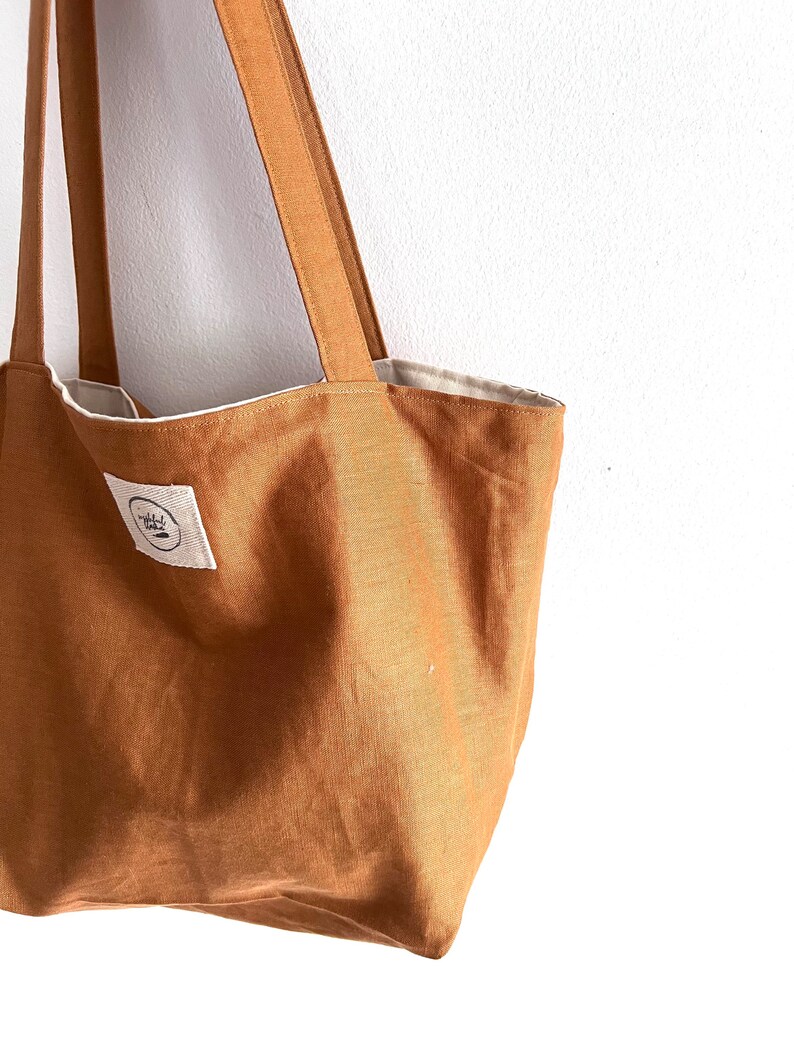 May include: A solid tobacco-colored tote bag featuring two long shoulder straps and a white rectangular fabric label on the front. The label contains a circular logo with black text and a small graphic element. The interior lining is off-white.