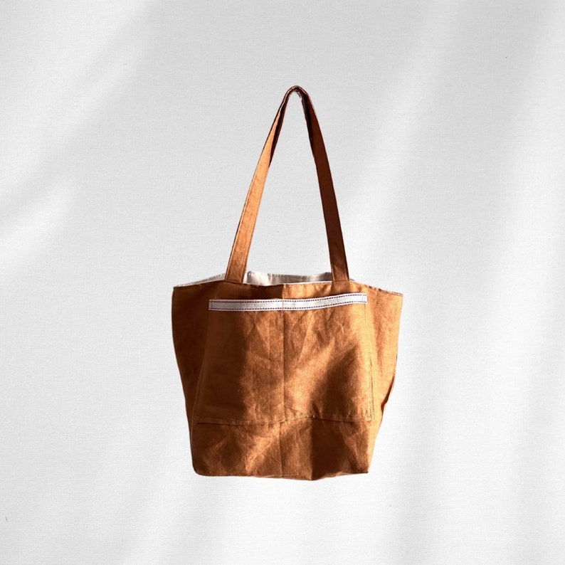 May include: A medium-brown tote bag featuring two long shoulder straps and a large exterior pocket on the front. The pocket is accented with a horizontal white stitched trim along the top edge. The bag has a structured base and a simple design.