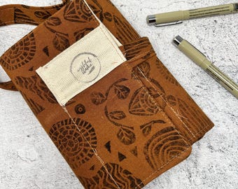 May include: A brown fabric pen case with a repeating pattern of abstract shapes and leaves. The case has a cream-colored pocket with the text "insightful wishes". Two beige pens with silver tips are next to the case.