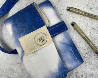 May include: A blue and white tie-dye pen roll with a matching tie. The roll is made of fabric and has a beige label with black text. Two pens are visible next to the roll.