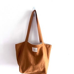 May include: A solid tobacco-colored tote bag with two shoulder straps. The bag features a rectangular white fabric label stitched to the front center, displaying the circular text logo "mindful linen" in black ink. The bag has a simple, open design.
