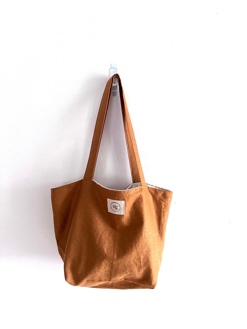 May include: A solid tobacco-colored tote bag with two shoulder straps. The bag features a rectangular white fabric label stitched to the front center, displaying the circular text logo "mindful linen" in black ink. The bag has a simple, open design.