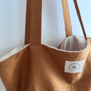 May include: A tan tote bag with two long shoulder straps and a white interior lining. A small, rectangular white fabric patch is sewn onto the front, featuring a circular logo with indistinct black text and a horizontal line graphic.