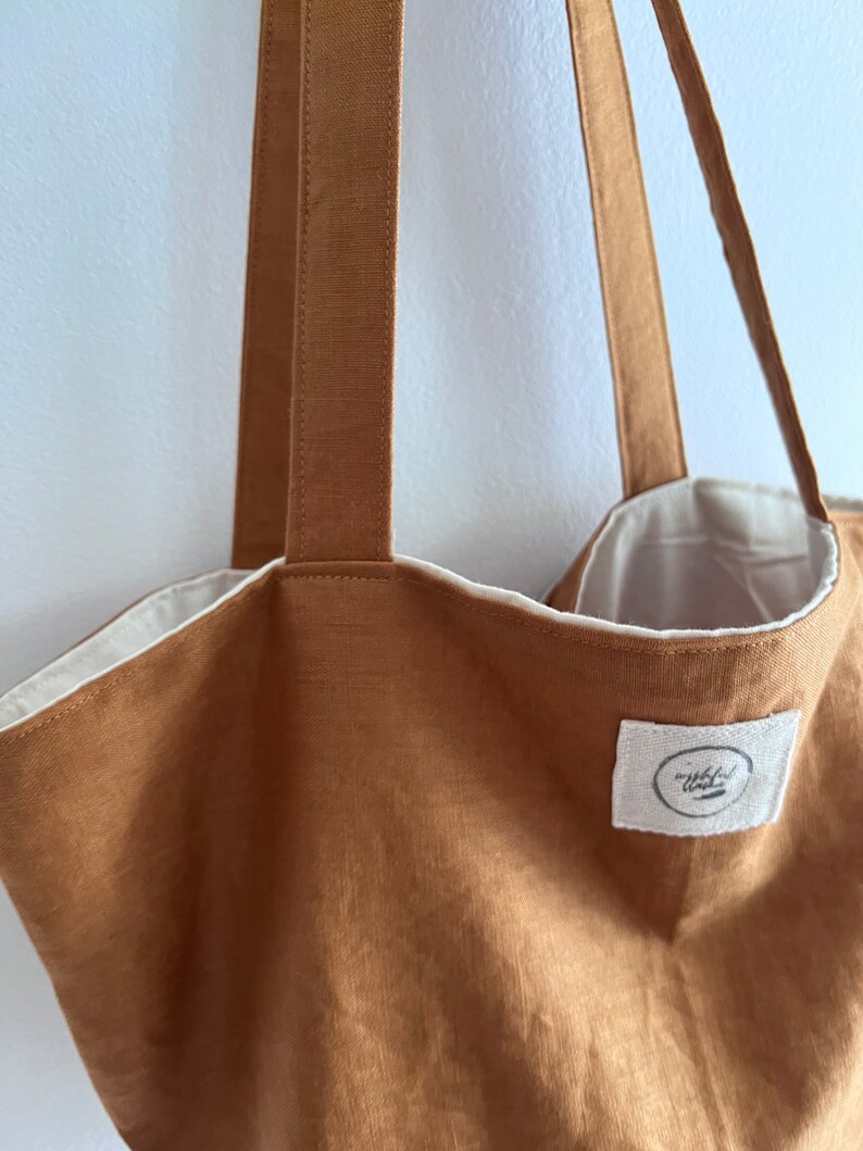 May include: A tan tote bag with two long shoulder straps and a white interior lining. A small, rectangular white fabric patch is sewn onto the front, featuring a circular logo with indistinct black text and a horizontal line graphic.
