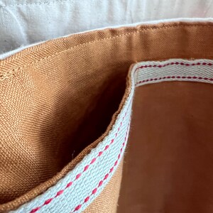 May include: The interior of a tan fabric tote bag featuring a light-colored woven trim along the top edge. The trim is accented with a thin, horizontal red stitched line. The bag has a visible seam construction and a sturdy, textured fabric lining.