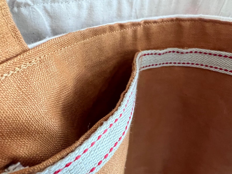 May include: The interior of a tan fabric tote bag featuring a light-colored woven trim along the top edge. The trim is accented with a thin, horizontal red stitched line. The bag has a visible seam construction and a sturdy, textured fabric lining.
