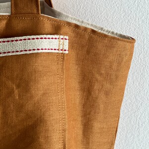 May include: A solid tobacco-colored tote bag made of a textured fabric. The bag features a cream-colored strap detail across the front, accented with two horizontal rows of red stitching. The interior lining is a light cream-colored fabric.
