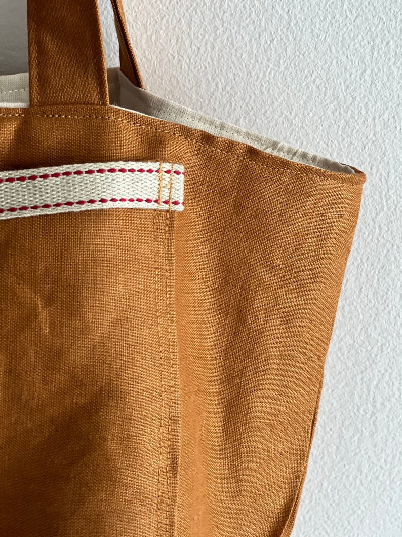 May include: A solid tobacco-colored tote bag made of a textured fabric. The bag features a cream-colored strap detail across the front, accented with two horizontal rows of red stitching. The interior lining is a light cream-colored fabric.