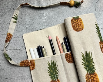 May include: A beige canvas pencil roll with a pineapple print. The roll has a brown strap with a wooden bead. The roll is unrolled and shows 10 pencils and pens inside.