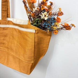 May include: A mustard yellow tote bag with a cream-colored interior lining and two exterior pockets featuring a cream trim with red stitching. The bag is filled with a bouquet of artificial flowers in shades of orange, blue, and white.
