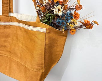 May include: A mustard yellow tote bag with a cream-colored interior lining and two exterior pockets featuring a cream trim with red stitching. The bag is filled with a bouquet of artificial flowers in shades of orange, blue, and white.