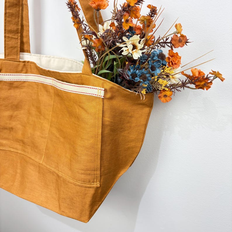 May include: A mustard yellow tote bag with a cream-colored interior lining and two exterior pockets featuring a cream trim with red stitching. The bag is filled with a bouquet of artificial flowers in shades of orange, blue, and white.