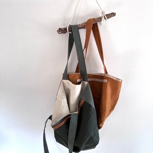 May include: Two tote bags hanging from a wooden branch. One bag is dark olive green with a light beige interior lining and long shoulder straps. The second bag is a solid tan color with a visible horizontal seam and matching tan shoulder straps.