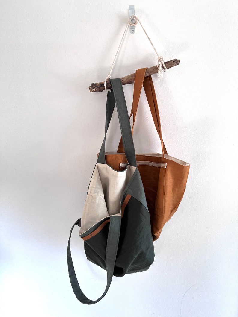 May include: Two tote bags hanging from a wooden branch. One bag is dark olive green with a light beige interior lining and long shoulder straps. The second bag is a solid tan color with a visible horizontal seam and matching tan shoulder straps.