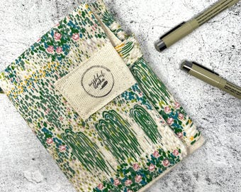 May include: A fabric notebook cover featuring a pattern of green weeping willow trees and small pink flowers on a cream background. A rectangular canvas label with the text Wishful Willow is stitched to the front. Includes a matching fabric bookmark.