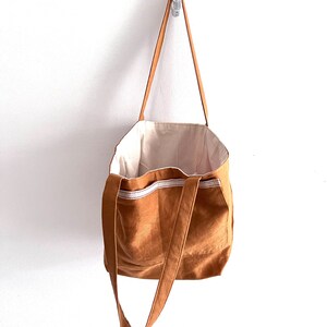May include: A tan tote bag with long shoulder straps and a cream-colored interior lining. The exterior features a horizontal band of white fabric with a thin red stitched line detail across the upper portion of the bag, which is hanging vertically.