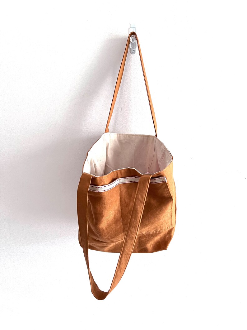 May include: A tan tote bag with long shoulder straps and a cream-colored interior lining. The exterior features a horizontal band of white fabric with a thin red stitched line detail across the upper portion of the bag, which is hanging vertically.
